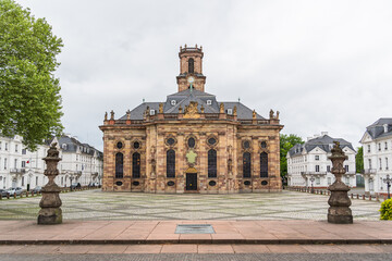 A large building Baroque church in the city of Saarbrucken in Germany with a clock tower and a small courtyard in front of it