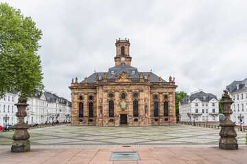 A large building Baroque church in the city of Saarbrucken in Germany with a clock tower and a small courtyard in front of it