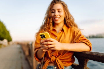 A young woman stands with a phone in her hands walking along the embankment at sunset. A beautiful...