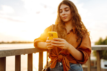 A young woman stands with a phone in her hands walking along the embankment at sunset. A beautiful...