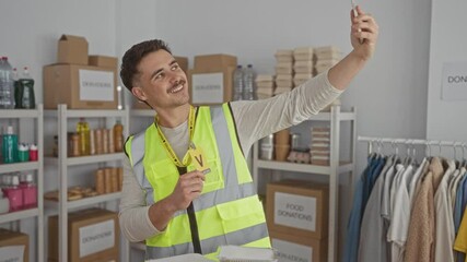Young hispanic man in reflective vest takes a selfie at an indoor charity center filled with donations, surrounded by organized boxes and clothing racks.