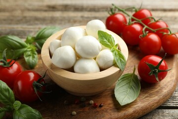 Tasty mozzarella cheese balls in bowl, basil, peppercorns and tomatoes on wooden table, closeup