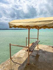 Rusty wooden bench with canopy on concrete pier by metal fence overlooking sea, in cloudy weather, perfect for themes of isolation