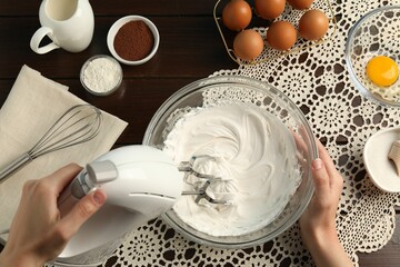 Woman whisking cream with hand mixer at wooden table, top view