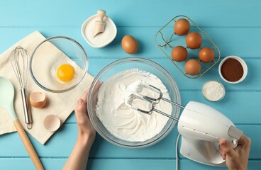 Woman whisking cream with hand mixer at light blue wooden table, top view