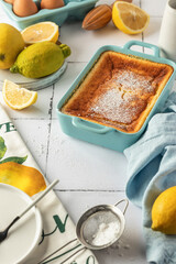 Baked lemon pudding with lemon zest and juice garnished with powdered sugar in blue baking dish on white tile background
