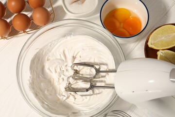 Hand mixer with whipped cream, eggs and other products on white wooden table, flat lay