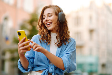 Beautiful woman with curly hair wearing headphones and using a mobile phone is having fun on a city street. Young woman enjoys listening to music and watching sunset outdoors. Tourism concept.