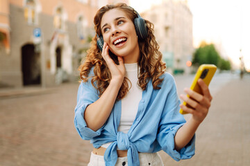 Beautiful woman with curly hair wearing headphones and using a mobile phone is having fun on a city...