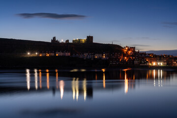 Fototapeta premium Whitby's historic skyline at dusk, with the illuminated Abbey ruins atop the headland, and the town's lights shimmering on the tranquil harbour waters below