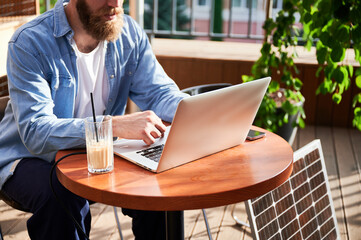 Cropped view of man works on laptop at outdoor round table, with glass of iced coffee. Solar panel nearby, emphasizing modern eco-friendly workspace that combines technology and sustainability.