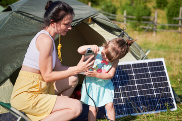 Young child and mother play with smartphone while charging with photovoltaic solar panel near tourist tent in summer. Integration of renewable energy in outdoor camping activities.