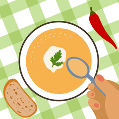 Pumpkin soup with sour cream and parsley. Someone is eating a tasty dish with a spoon. A plate with a delicacy on a tablecloth. A piece of bread and chili pepper