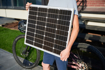 Close up view of person holding solar panel for charging, stands in front of brick building. Next to him pink electric mountain bike with black helmet resting on handlebars. Cropped view.