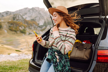 Naklejka premium Beautiful woman in the trunk of a car with a phone enjoying a road trip. Young traveler blogging and observing the mountain scenery from the car. Wanderlust.