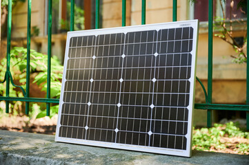 Photovoltaic solar panel positioned against green metal fence, capturing sunlight in residential garden. Lush vegetation and house in background, highlighting sustainable living in urban environment.