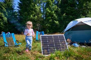 Young child playing near tourist tent with photovoltaic solar panel in summer. On background of dense forest. Integration of renewable energy in outdoor camping activities.