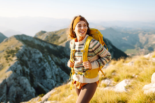 A young female traveler with a colorful hiking backpack walks along a mountain peak. A cheerful woman feels free and enjoys the mountain landscape at sunset. Hiking concept. Active lifestyle.
