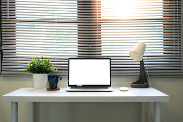 Bright home office with a laptop, coffee cup, indoor plants, and lamp under window blinds
