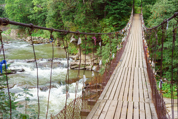 Wooden suspension bridge with colorful ribbons hanging from wires crosses fast mountain river, mountain creek in lush green coniferous Carpathian forest. Adventure, travel, hiking and local charm.