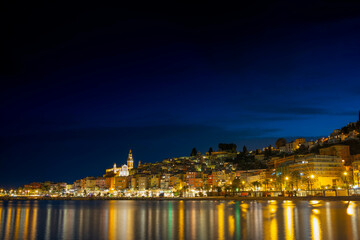 Night panorama of Menton in France. The lights of the night city are reflected on the calm sea surface.