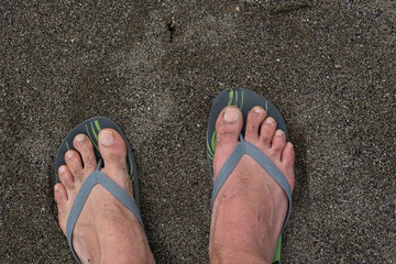 Male feet in rubber flip-flops standing on sand. Feet in rubber flip-flops standing on sandy beach,...