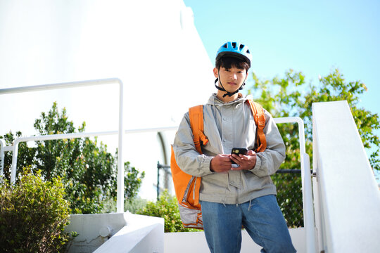 Young Asian delivery guy standing along the stairs while holding his phone