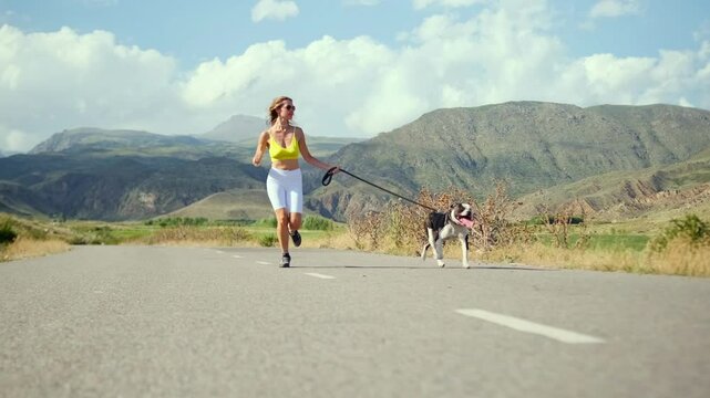 A beautiful, fit woman in a yellow crop top and white bike shorts runs with pit bull along a road, surrounded by lush landscapes and towering mountains, perfect for an energizing workout.
