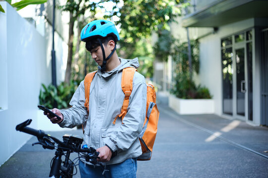 Young Asian delivery guy holding his bicycle while checking his phone