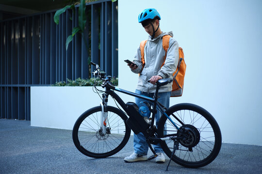 Young Asian delivery guy standing beside his bicycle while on his phone.