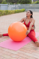 Latin woman doing pilates exercises with orange fitness ball outdoors