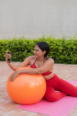 Latin woman doing pilates with fitness ball and taking selfie with smartphone