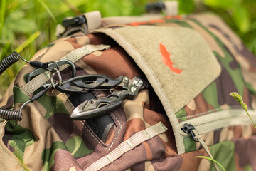 Fishing bag. Defocus military backpack with scissors. Army bag on green grass background. Military camouflage army rucksack. Tourist summer hiker. Sunny. Close-up. Outdoor. Out of focus. 