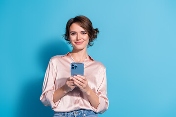 Stylish young woman in satin pink blouse uses smartphone against blue background