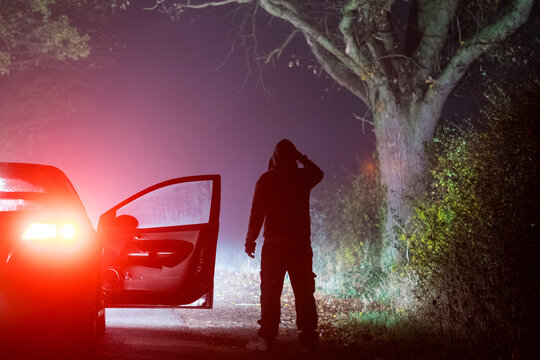 A horror concept of a spooky hooded figure, standing by a parked car. On an eerie lonely foggy road at night in the countryside.