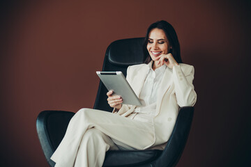 Successful businesswoman in stylish white suit using a tablet while relaxed in a modern chair...