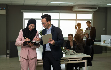 Professional office environment with colleagues collaborating. Businessman using digital tablet and woman in hijab taking notes while other team members working in background in open plan