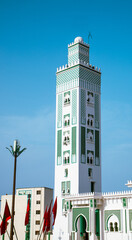 Obraz premium Elegant white and green minaret of a mosque in Tetouan, Morocco, under a clear blue sky, surrounded by red Moroccan flags.