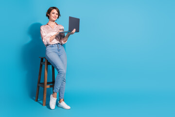 Trendy young woman in pink satin blouse holding laptop against blue background while sitting...