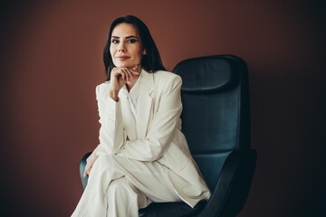 Young businesswoman in a white suit sitting confidently on a chair against a simple brown background