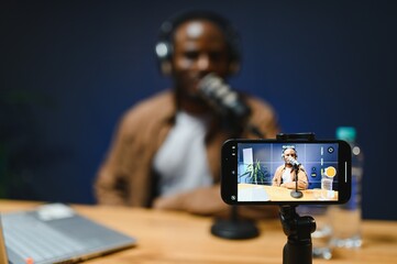 Black vlogger filming podcast on smartphone in studio. Cheerful blogger