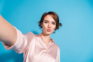 Charming young woman in satin pink blouse taking a selfie with a playful expression against a...