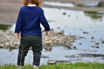 Red-haired toddler girl in dark clothes stands near puddles and rocky ground. Scene gives a moody,...