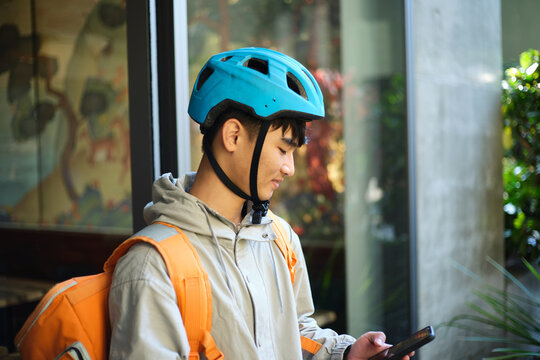 Asian delivery guy looking on his phone outside restaurant wearing bike helmet