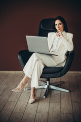 Beautiful businesswoman in a white suit seated in a modern office chair with a laptop on a...