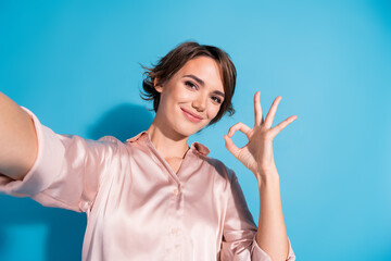 Charming young woman showing okay gesture against blue background in stylish pink satin blouse