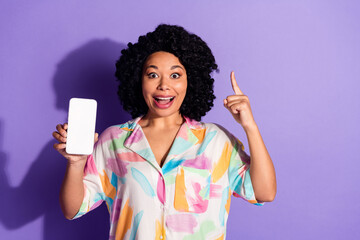 Excited woman holding smartphone and pointing up against vibrant violet background in colorful...