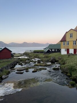 Evening light over Gj&oacute;gv village, Eysturoy, Faroe Islands. A quiet stream winds between turf-roof houses and flows into the ocean, framed by distant mountains.