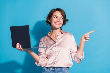 Young woman in satin blouse holding laptop against blue background while pointing and smiling in a...