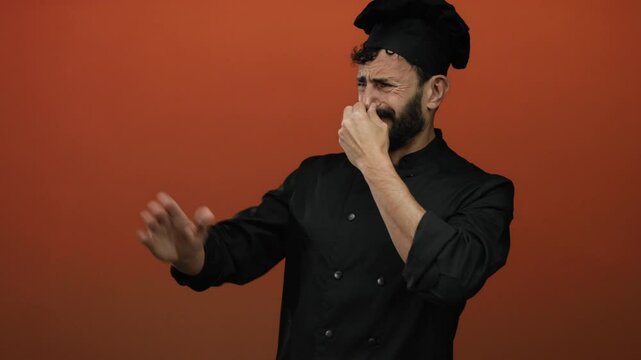 Hispanic male chef in black uniform grimacing against red background, indicating bad smell.
